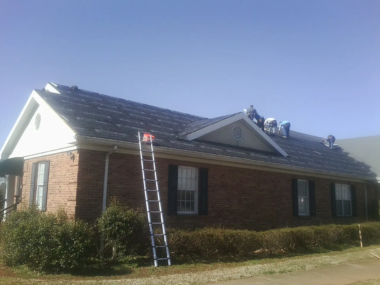 Crew tearing shingles off a brick ranch roof with a ladder set — Emergency Roof Repair work by our crew in Saranac