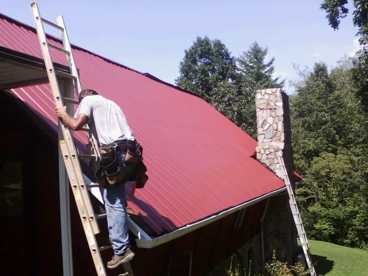 Roofer on a ladder beside a red metal roof and stone chimney — Soffit & Fascia Repair by Roofing Brothers in Saranac