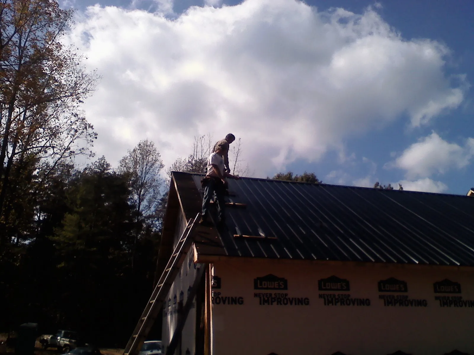 Roofer fastening dark metal panels high against a cloudy sky, from a Roof Inspection job in Saranac